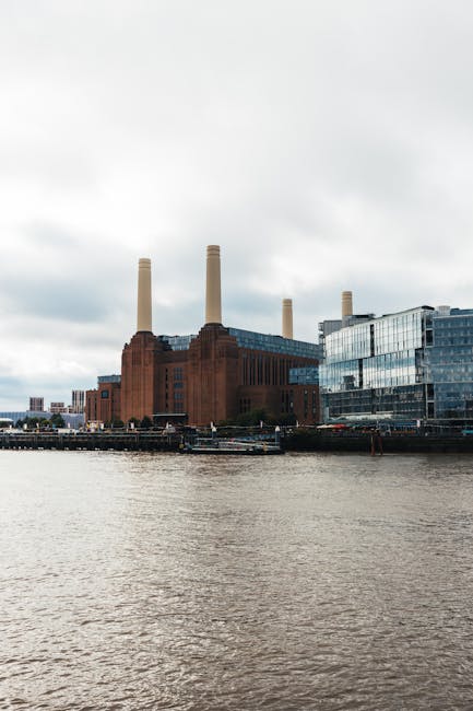 A low-angle view of the Battersea Power Station showing two tall, white, cylindrical chimneys with vertical ridges, set against a bright blue sky with a few scattered clouds. The base of the building 