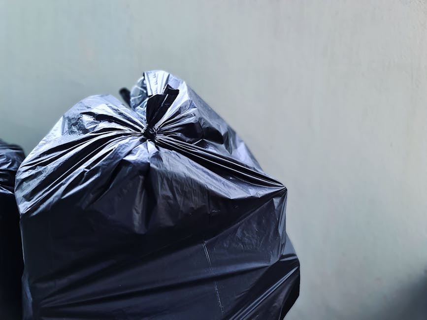Close-up of two black plastic rubbish bags tightly knotted at the top, placed against a plain, light-colored wall. The bags are made of shiny, smooth plastic material, with visible creases and folds o
